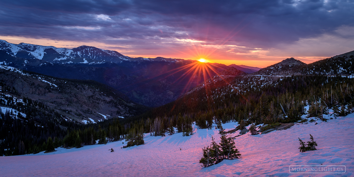 The sun breaks through the clouds at tree line on an early June morning while snow still clings to the sides of the mountain...